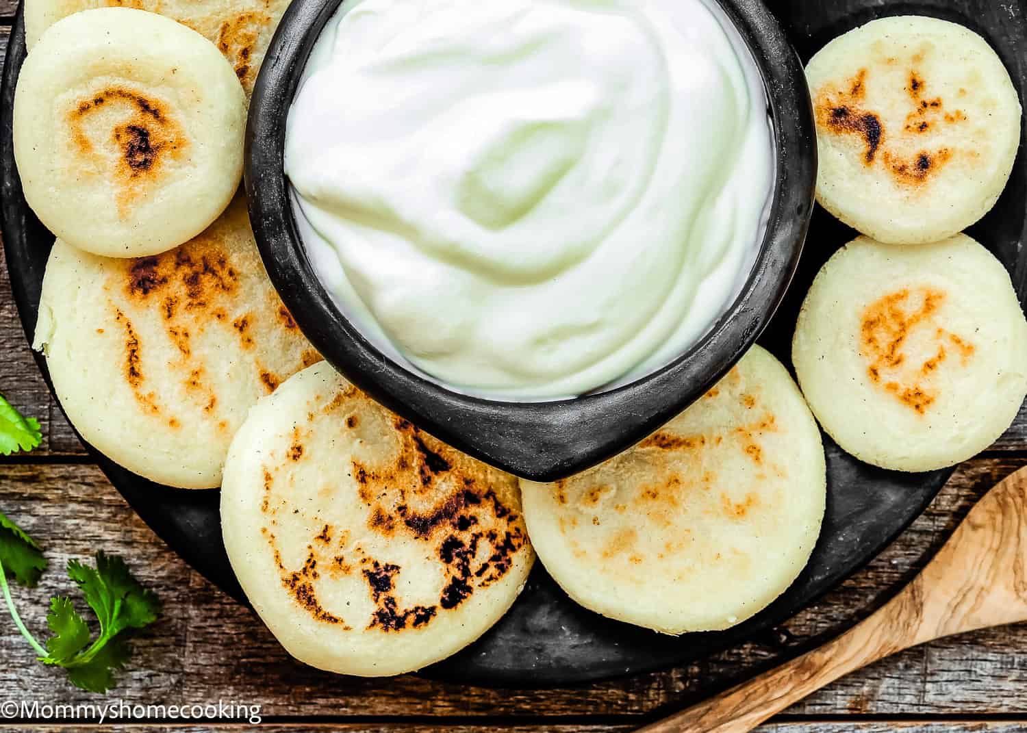 A plate of grilled arepas surrounds a bowl of creamy white dip, set on a wooden surface with a wooden spoon and some cilantro—perfect for showcasing How to Make Venezuelan Arepas (Arepas Venezolanas).