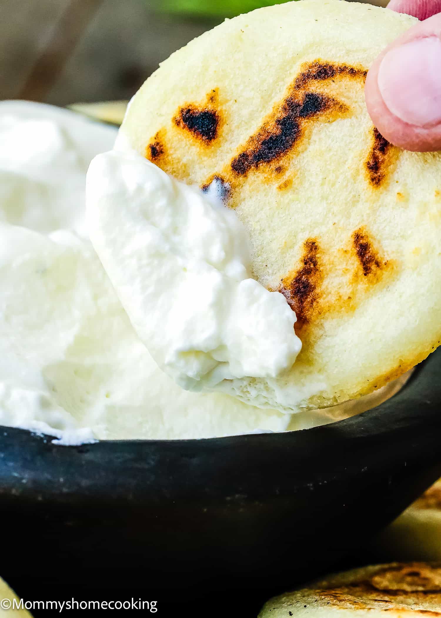 A hand dips a grilled arepa into a bowl of white cream, showcasing the delicious result of learning How to Make Venezuelan Arepas (Arepas Venezolanas).