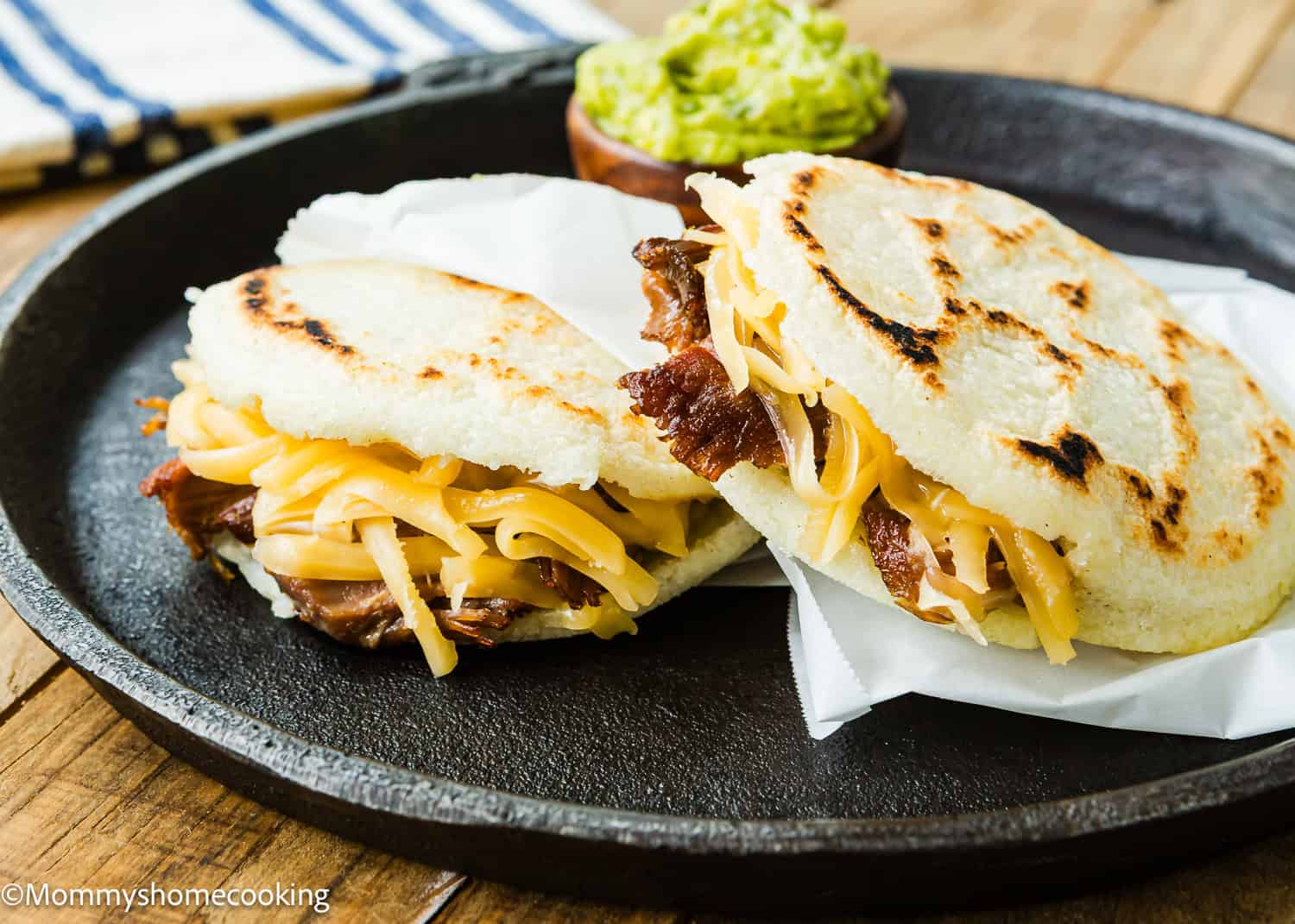 Two arepas filled with shredded cheese and meat are served on a black plate, with a small bowl of guacamole in the background—an inviting example of How to Make Venezuelan Arepas (Arepas Venezolanas).