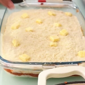 A glass baking dish filled with Easy Venezuelan Pasticho, layered and topped with grated cheese and small cubes of butter, ready to be baked.
