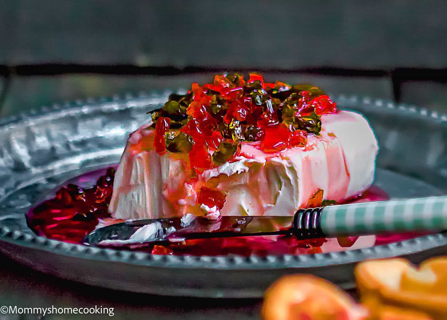A block of cream cheese topped with hot pepper jelly sits on a metal plate, making the perfect Hot Pepper Jelly Cream Cheese Appetizer (Sweet & Spicy!). A knife rests beside it for easy serving.