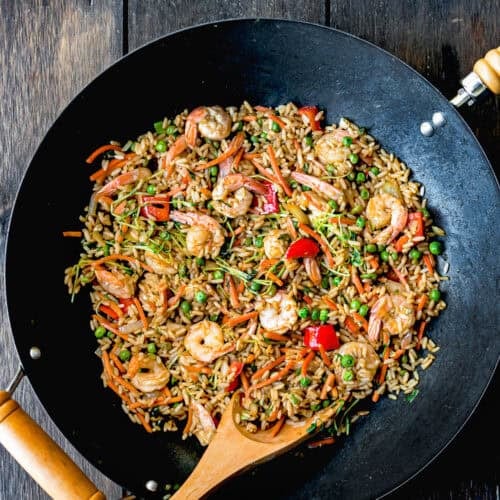 A wok brimming with easy shrimp fried rice, dotted with peas and vibrant red bell peppers, complemented by a wooden spoon resting on a dark wooden surface.