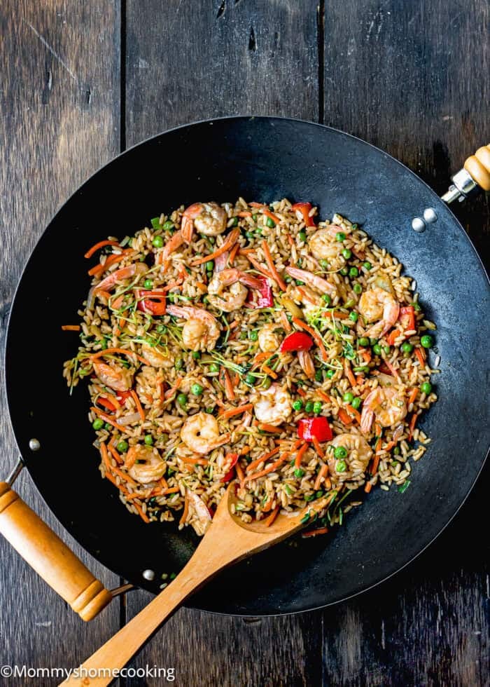 A wok brimming with easy shrimp fried rice, dotted with peas and vibrant red bell peppers, complemented by a wooden spoon resting on a dark wooden surface.