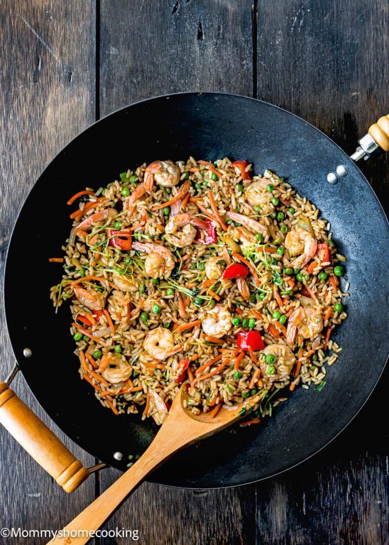 A wok brimming with easy shrimp fried rice, dotted with peas and vibrant red bell peppers, complemented by a wooden spoon resting on a dark wooden surface.