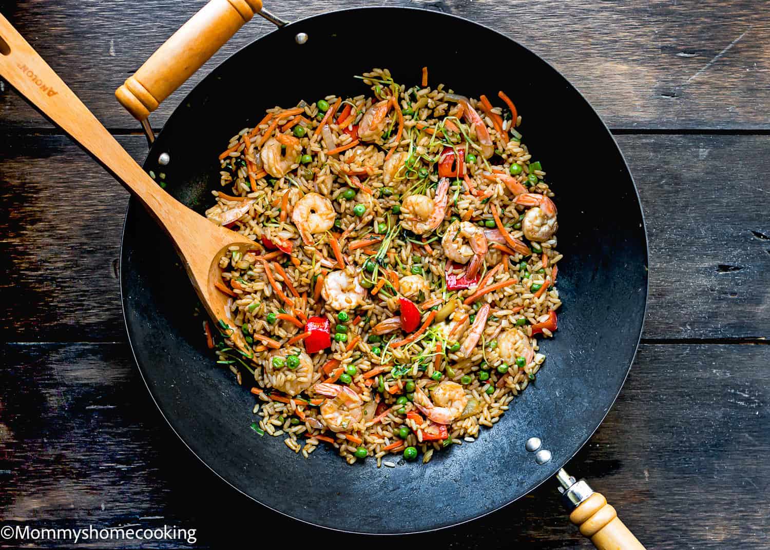 Easy Shrimp Fried Rice: Wok with shrimp fried rice, featuring peas, carrots, and red bell peppers, mixed with a wooden spoon on a dark wooden surface.