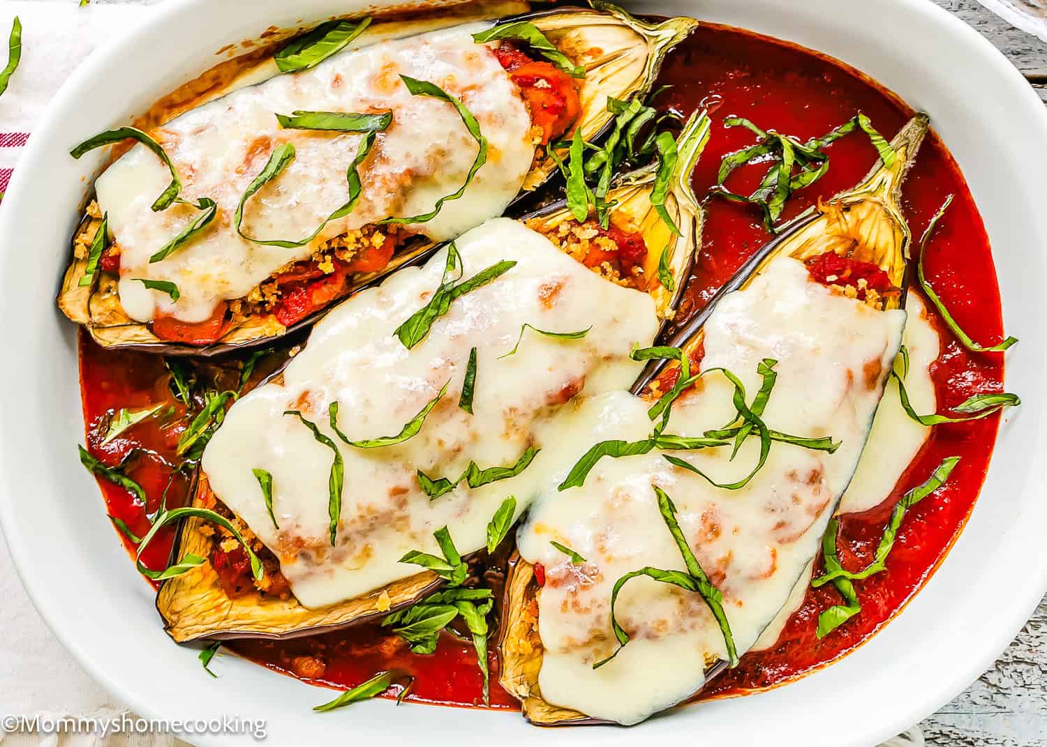 Three halved eggplants are transformed into Stuffed Eggplant Parmesan Boats, topped with tomato sauce, melted mozzarella cheese, and basil strips, then baked in a white dish filled with marinara sauce.