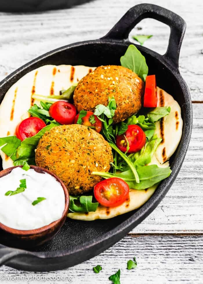 Quickly made falafel patties on flatbread feature alongside cherry tomatoes, arugula, and herbs. They're served in a skillet with a side of white sauce in a small wooden bowl.