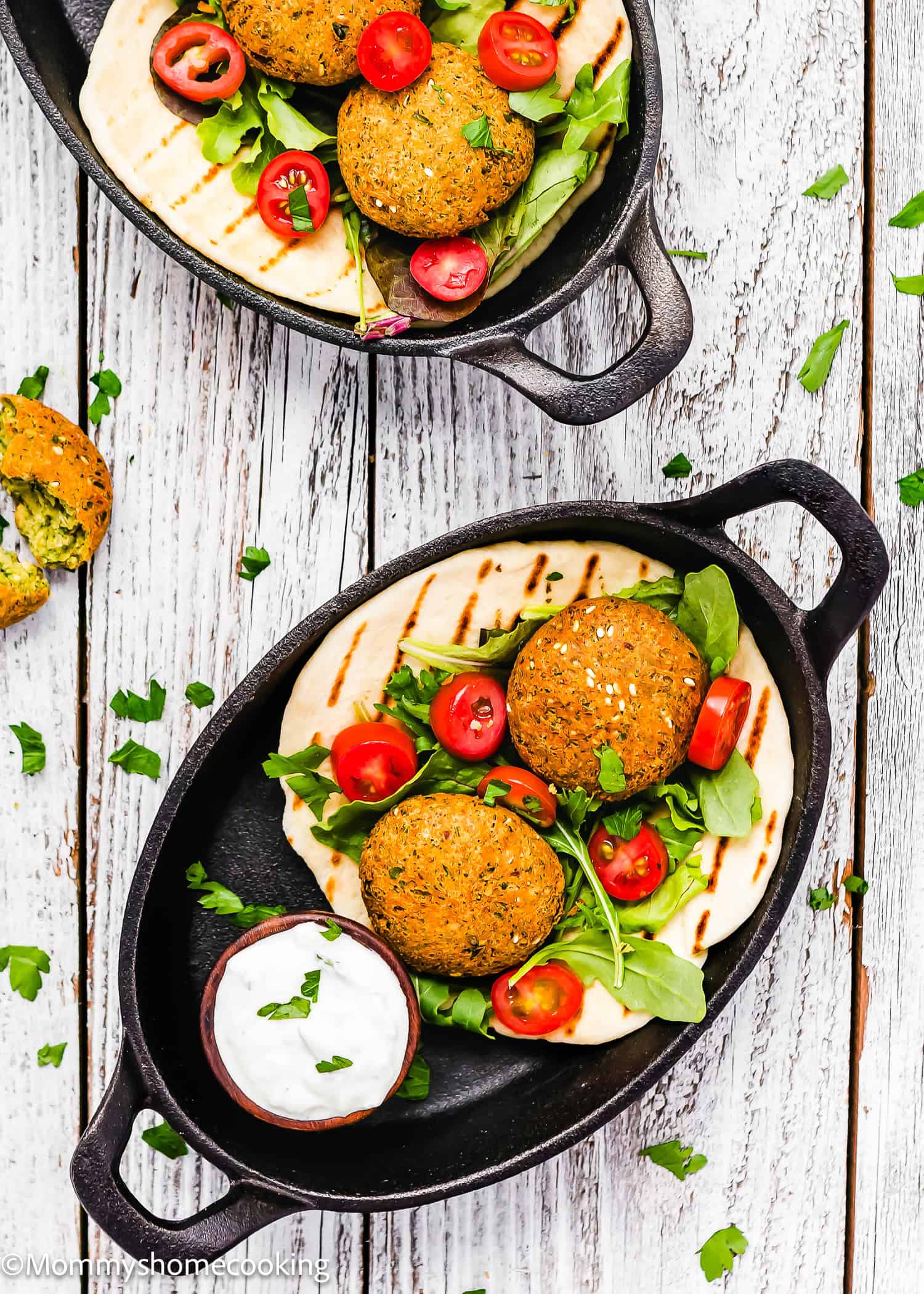 Two black dishes with falafel, sliced tomatoes, arugula, and pita surround a small bowl of yogurt sauce on a white wooden table. Discover how to make falafel the quick way for a delicious and speedy meal.