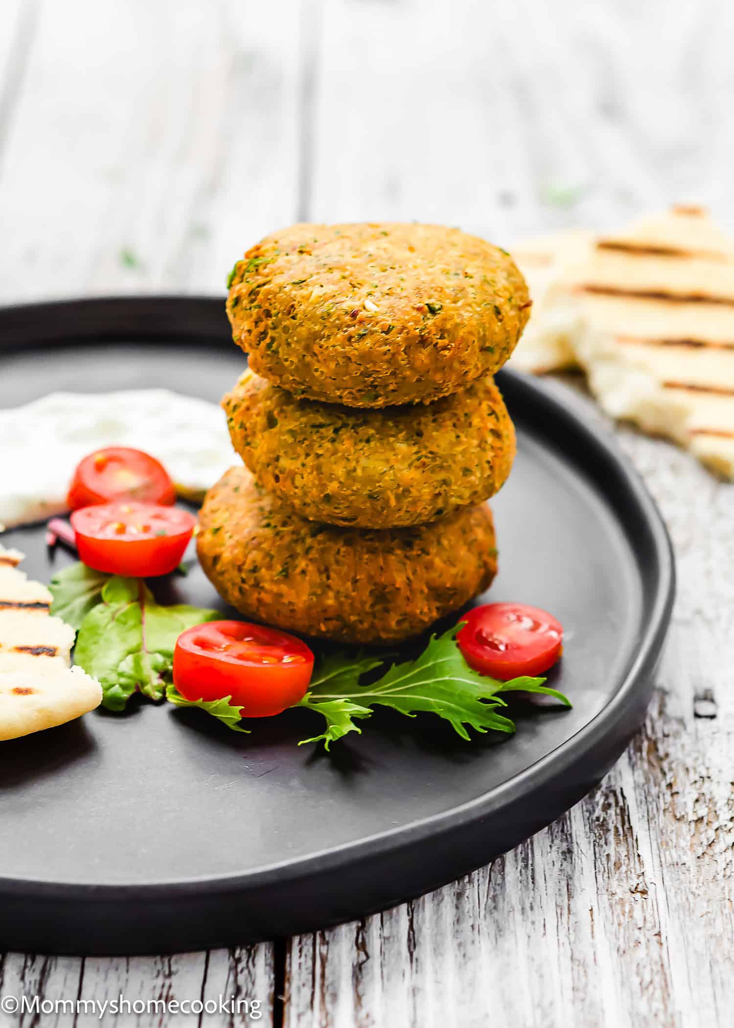 A trio of cooked falafel patties is arranged on a black plate, accompanied by vibrant sliced cherry tomatoes, grilled pita, and fresh greens, offering a glimpse into how to make falafel the quick way.