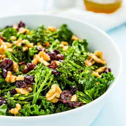 A white bowl containing a Chick-fil-A-inspired kale salad with walnuts, cranberries, and broccoli. Dressing is visible in the background.