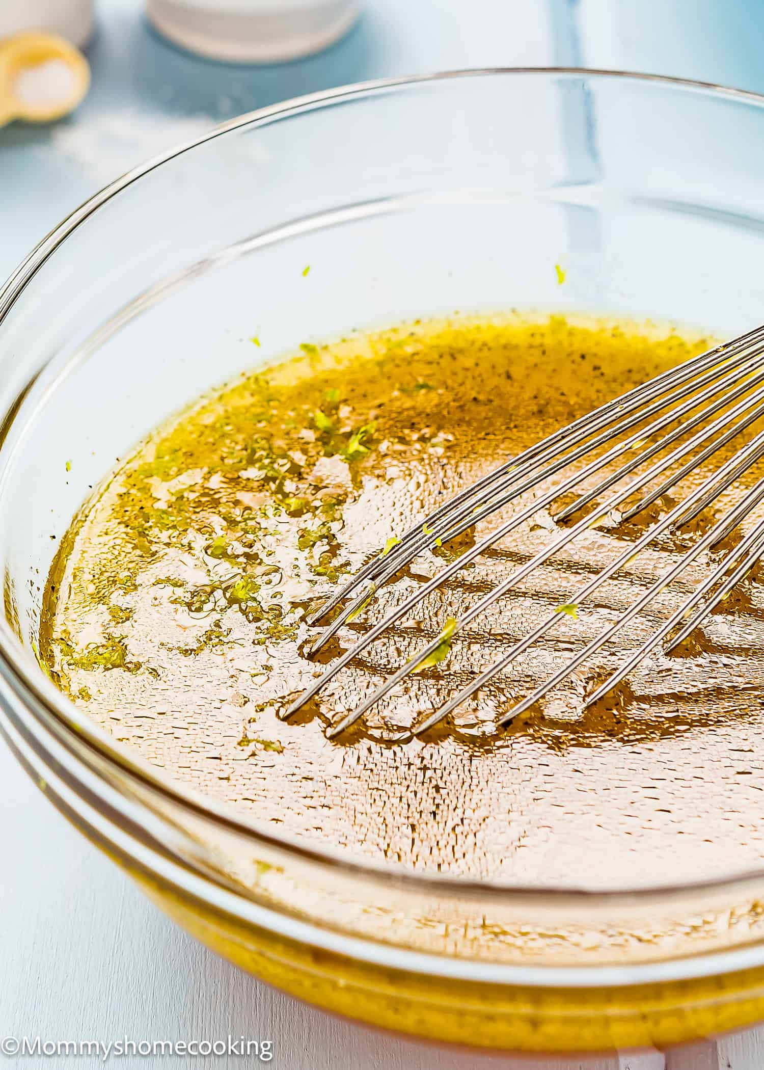 A glass bowl containing a yellowish liquid marinade, reminiscent of the dressing from a Chick fil A Kale Salad, is mixed with green herbs using a metal whisk.