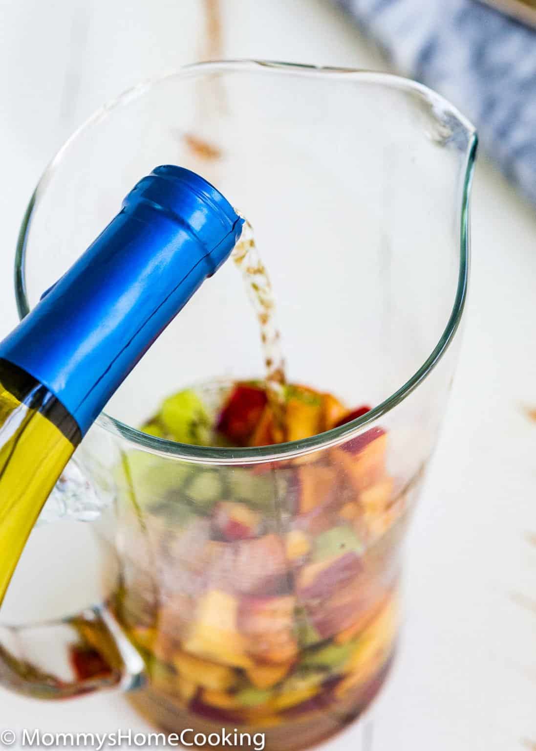A bottle of white wine is being poured into a glass pitcher filled with chopped fruit, creating an Easy Fruity White Sangria perfect for any gathering.