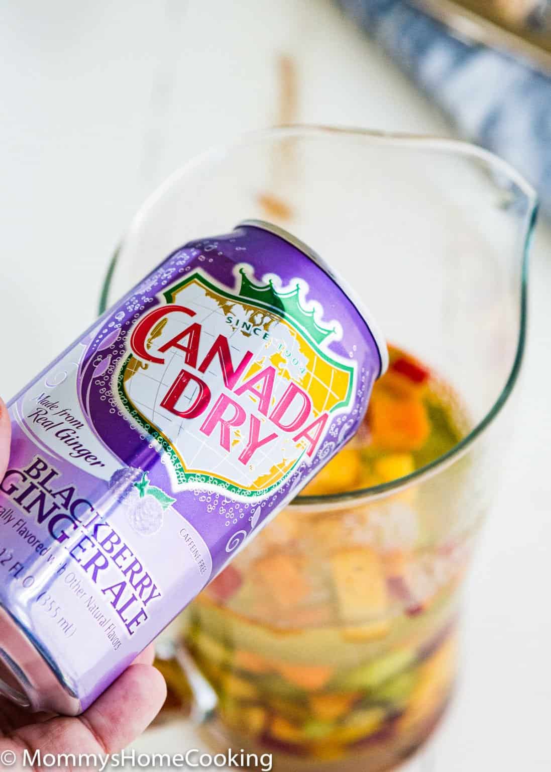 A hand holds a can of Canada Dry Blackberry Ginger Ale above a glass pitcher filled with fruit and liquid, making it perfect for an Easy Fruity White Sangria.