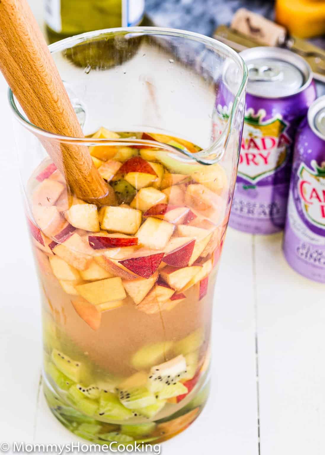 A glass pitcher filled with chopped apples, kiwi, and a light-colored drink—this Easy Fruity White Sangria is stirred with a wooden spoon. Purple Canada Dry cans are in the background, adding a playful touch.