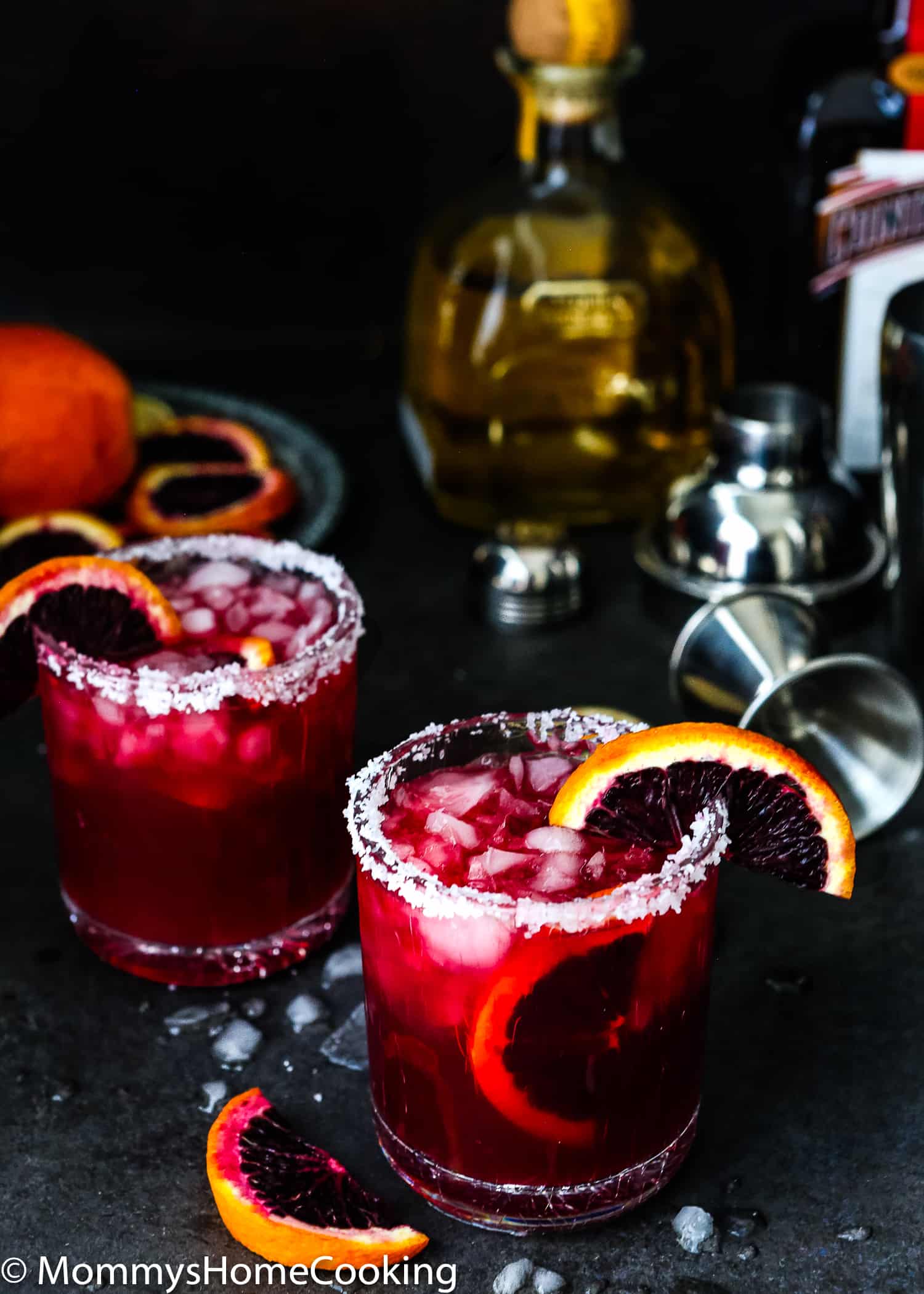 two blood orange margaritas in transparent glasses with a tequila bottle in the background and sliced oranges
