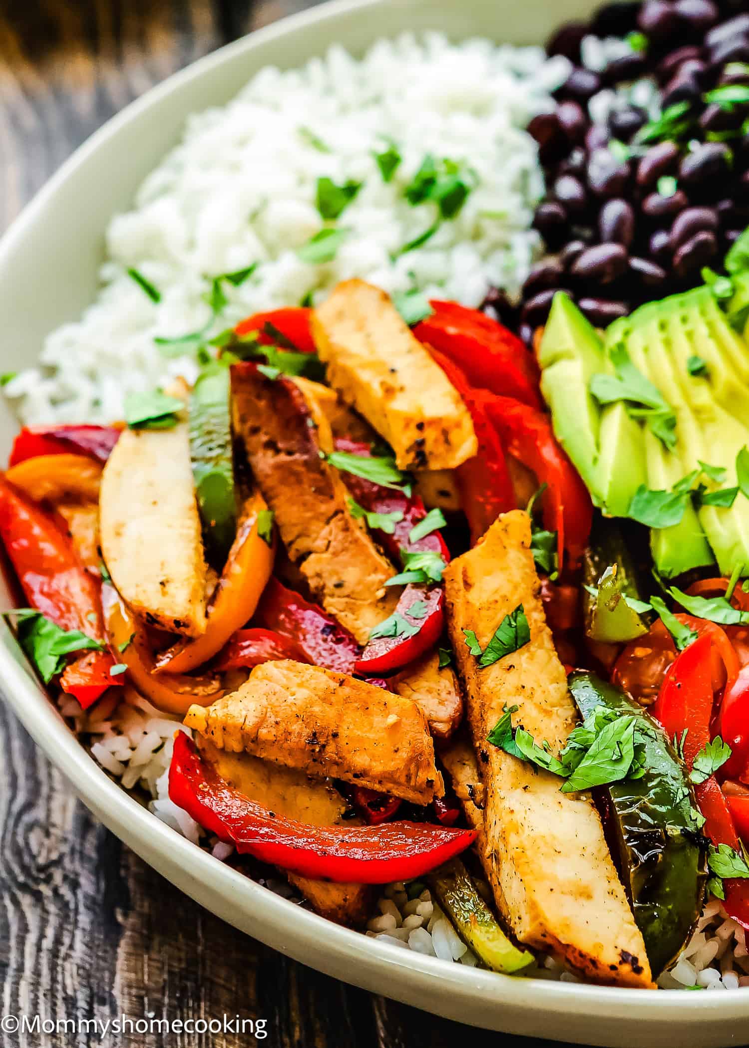 An easy turkey fajita bowl features rice, black beans, corn, cherry tomatoes, avocado slices, and seasoned grilled chicken strips. It's garnished with cilantro for a fresh touch.