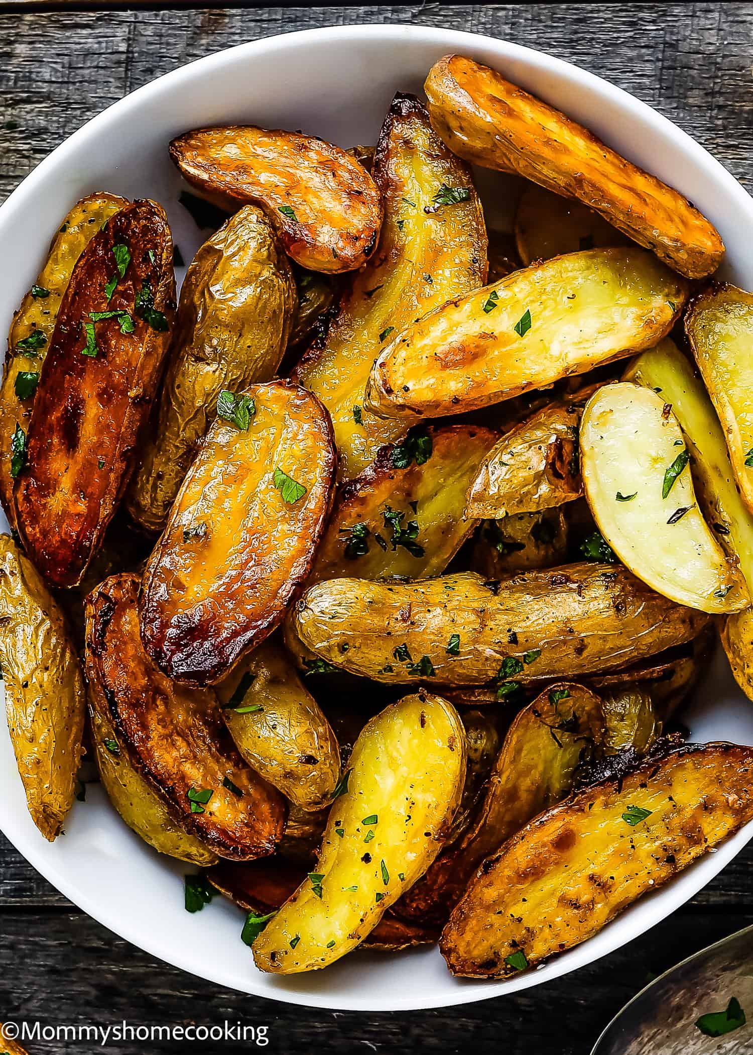 A bowl of easy garlic lemon roasted potatoes seasoned with herbs rests on a wooden surface.