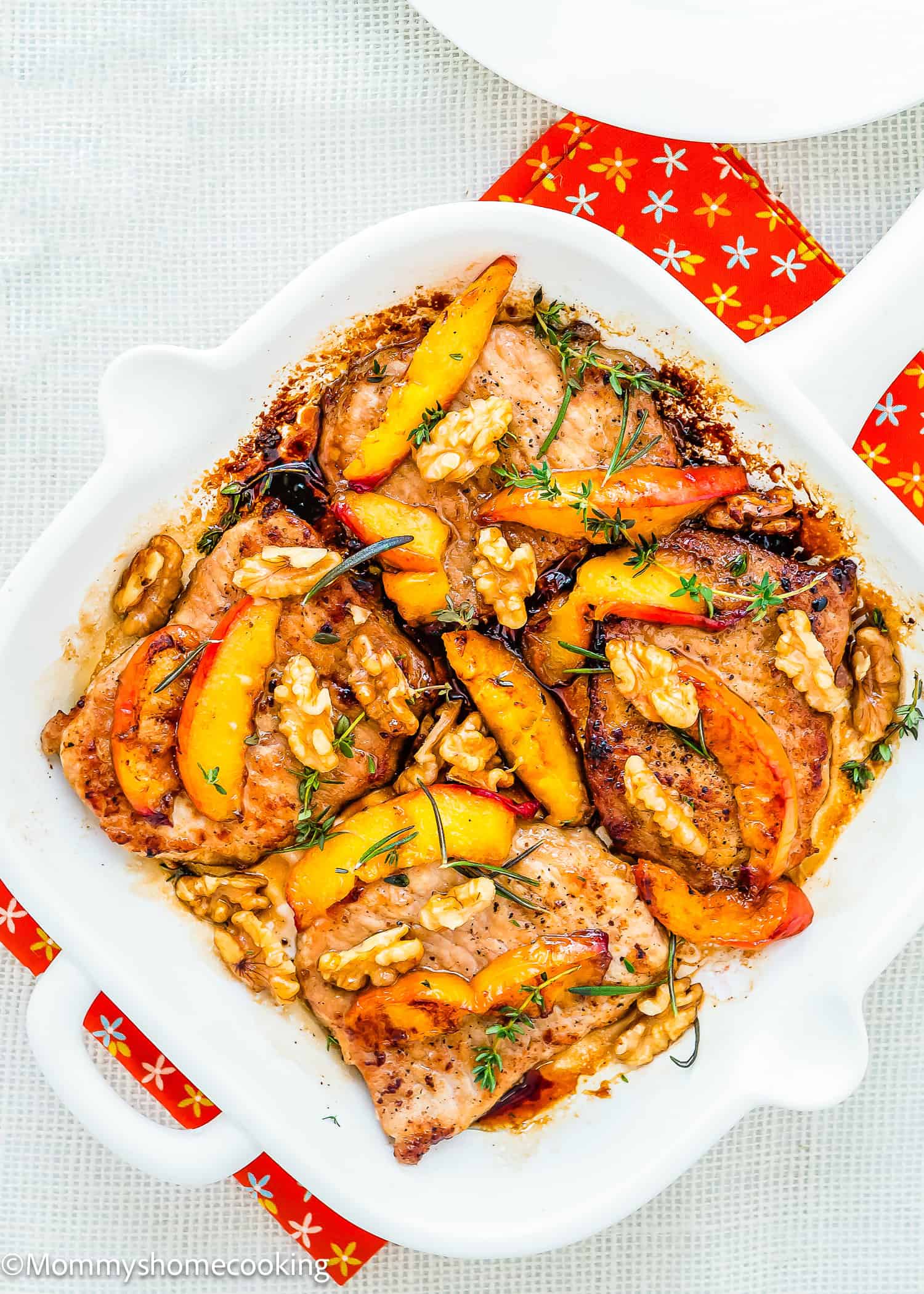 Grilled Pork Chops with Peaches in a white dish, topped with juicy peach slices, walnuts, and fresh herbs, served on a red patterned napkin.