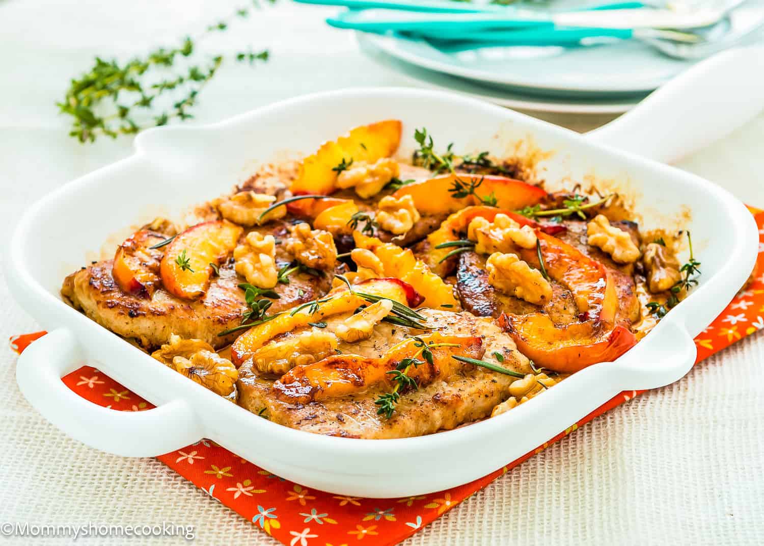 Baked chicken breasts topped with peach slices, walnuts, and fresh herbs share the table with Grilled Pork Chops with Peaches, all in a white ceramic baking dish on an orange placemat.
