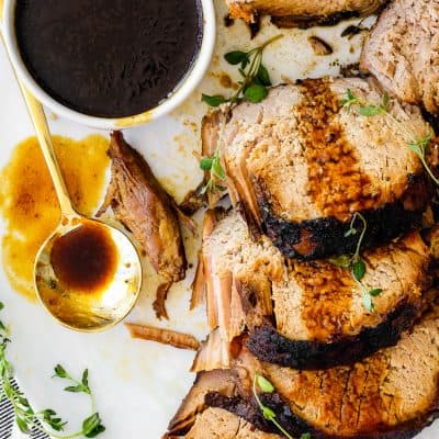 Sliced roast pork loin with gravy on a plate, garnished with herbs, sits beside a bowl of additional sauce. Perfectly tender from the slow cooker, this dish is a savory delight.