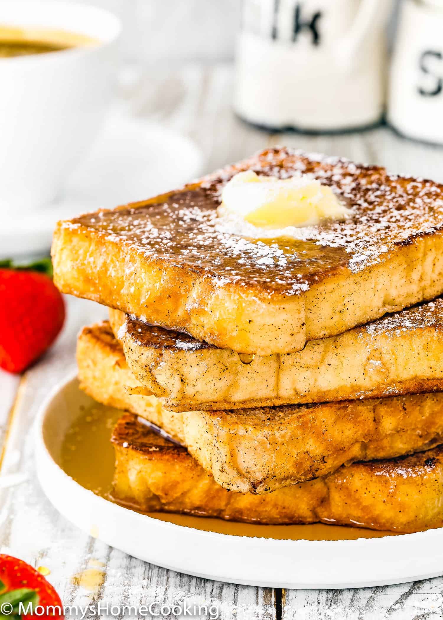 A stack of four eggless French toast in a plate with powdered sugar dusted on top and maple syrup.