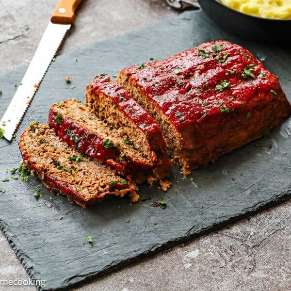 A sliced Egg-Free Meatloaf Recipe topped with ketchup glaze is served on a slate board with a serrated knife and bowls of mashed potatoes and chopped herbs nearby.