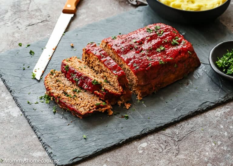 A sliced Egg-Free Meatloaf Recipe topped with ketchup glaze is served on a slate board with a serrated knife and bowls of mashed potatoes and chopped herbs nearby.