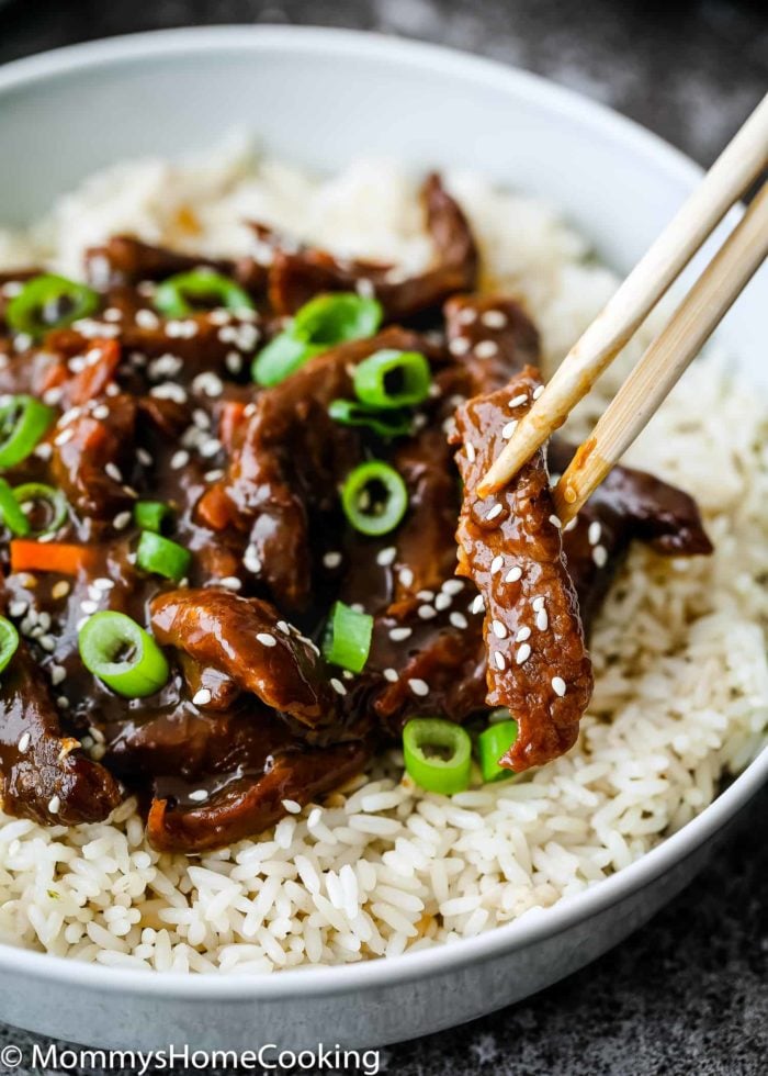 A bowl of Easy Instant Pot Mongolian Beef features saucy beef strips, chopped green onions, and sesame seeds atop a bed of rice. Chopsticks hold a piece of beef.