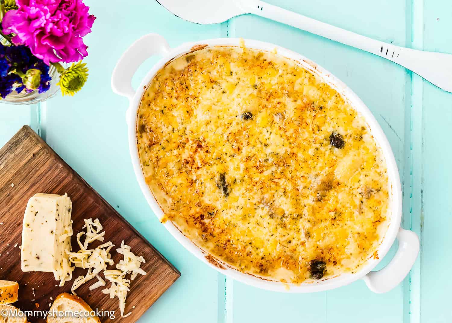 A Cheesy Gnocchi Casserole (Skillet Recipe) sits baked in an oval white dish on a blue surface, with shredded cheese, a cheese block, and croutons on a wooden board nearby, plus a vase of flowers in the corner.