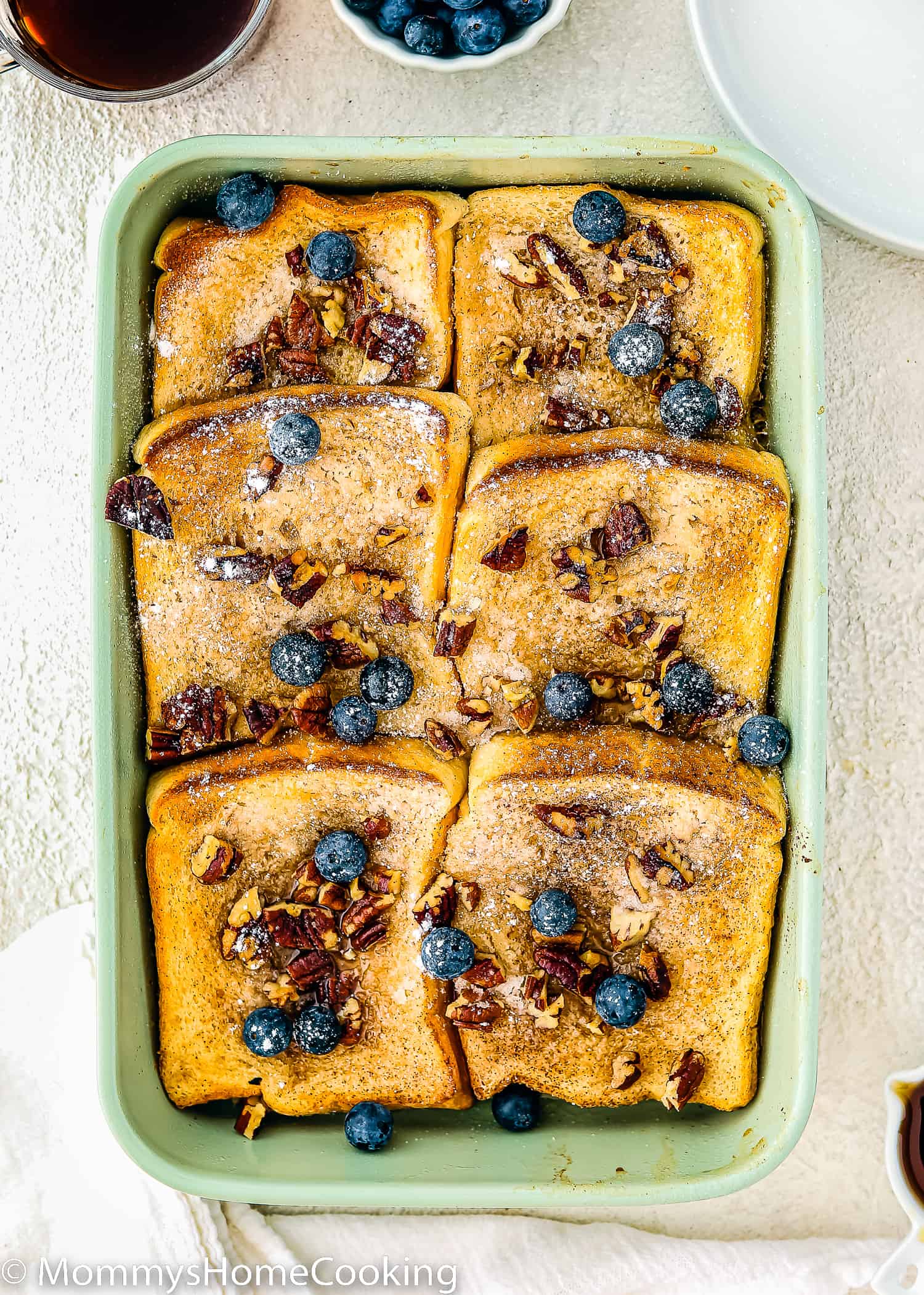 Eggless French Toast Casserole with powered sugar, pecan and blueberries on top over a white surface.