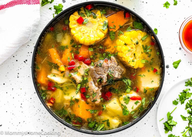 A bowl of Authentic Venezuelan Sancocho (Sancocho Venezolano) featuring beef, corn, carrots, potatoes, herbs, and red peppers in a clear broth, garnished with chopped cilantro.