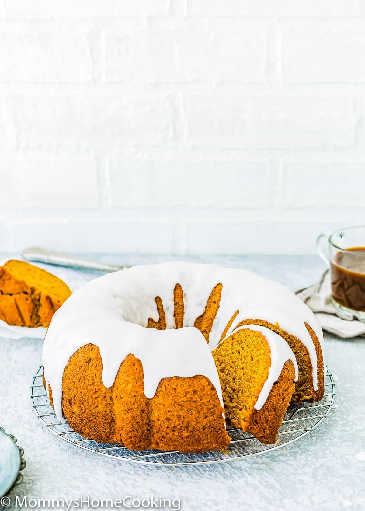 Sliced Eggless Pumpkin Cake with glaze over a cooling rack with a cup of coffee in the background .