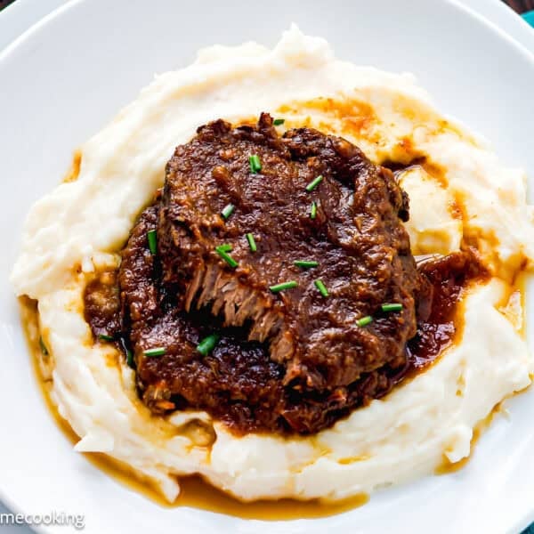 A serving of braised beef pot roast, inspired by a Venezuelan Asado Negro recipe, with rich gravy sits atop creamy mashed potatoes and is garnished with chopped chives on a white plate.