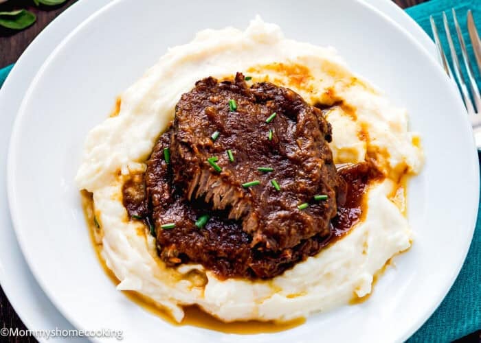 A serving of braised beef pot roast, inspired by a Venezuelan Asado Negro recipe, with rich gravy sits atop creamy mashed potatoes and is garnished with chopped chives on a white plate.