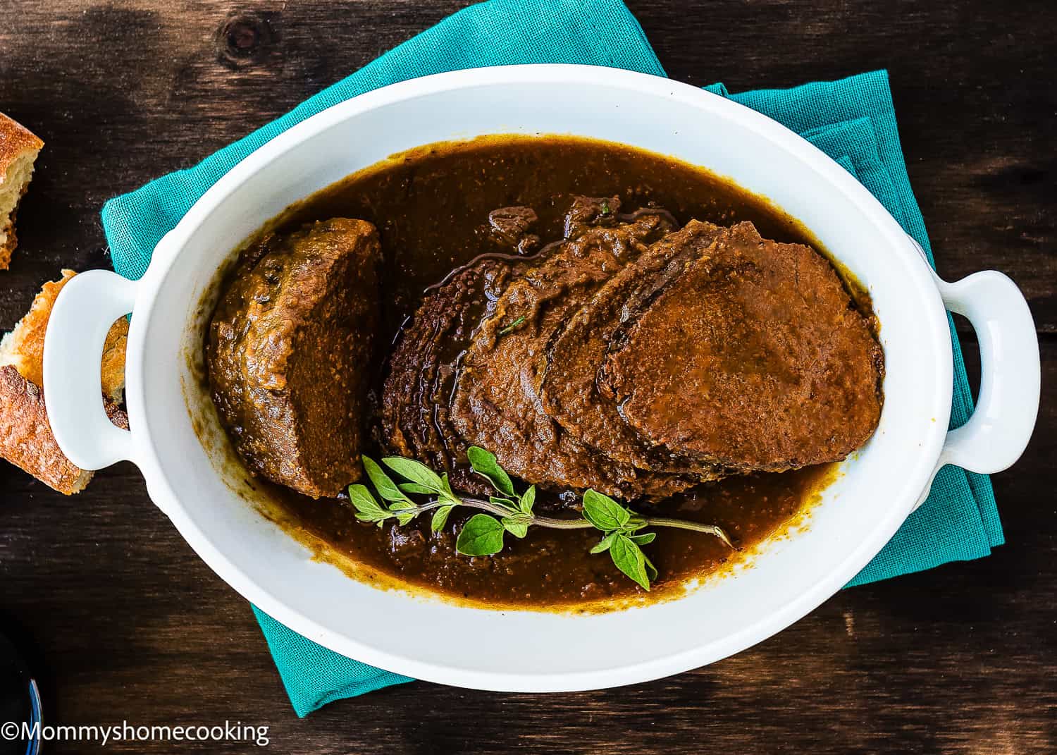 Sliced pot roast in rich brown gravy, inspired by a Venezuelan Asado Negro recipe, garnished with fresh herbs and served in a white oval dish on a teal cloth.
