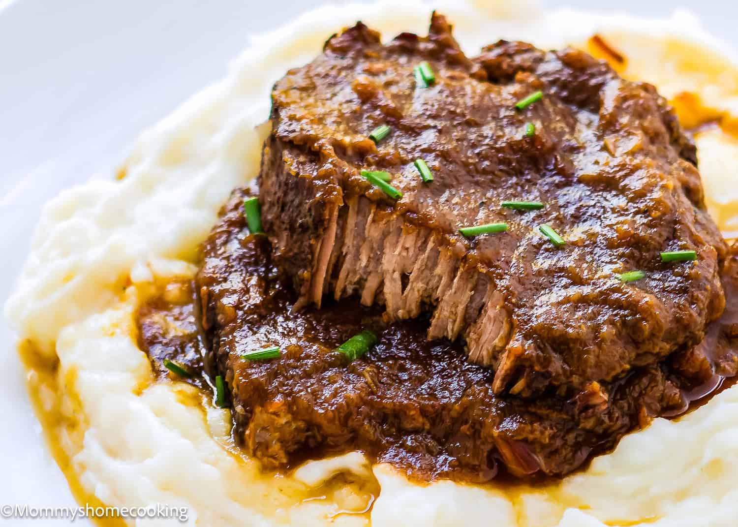 A slice of braised beef pot roast, inspired by the Venezuelan Asado Negro recipe, served on creamy mashed potatoes, topped with rich gravy and garnished with chopped chives.