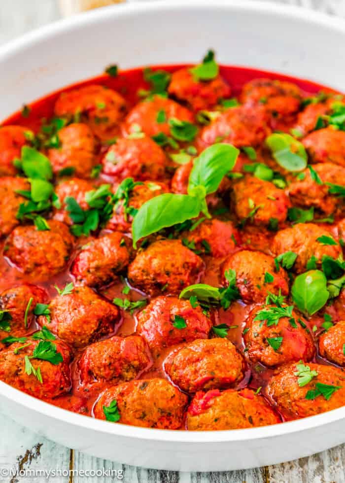 A white dish filled with the BEST Eggless Italian Meatballs in tomato sauce, garnished with fresh basil and parsley.