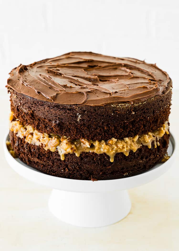 Two layers of egg-free chocolate cake on a cake stand filled with eggless coconut pecan filling and covered with chocolate frosting.