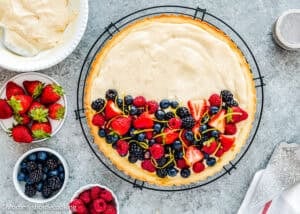 A partially decorated Easy Eggless Fruit Tart with Vanilla Pastry Cream, topped with assorted fresh berries and citrus zest, sits on a cooling rack. Bowls of strawberries, raspberries, and blackberries are nearby.
