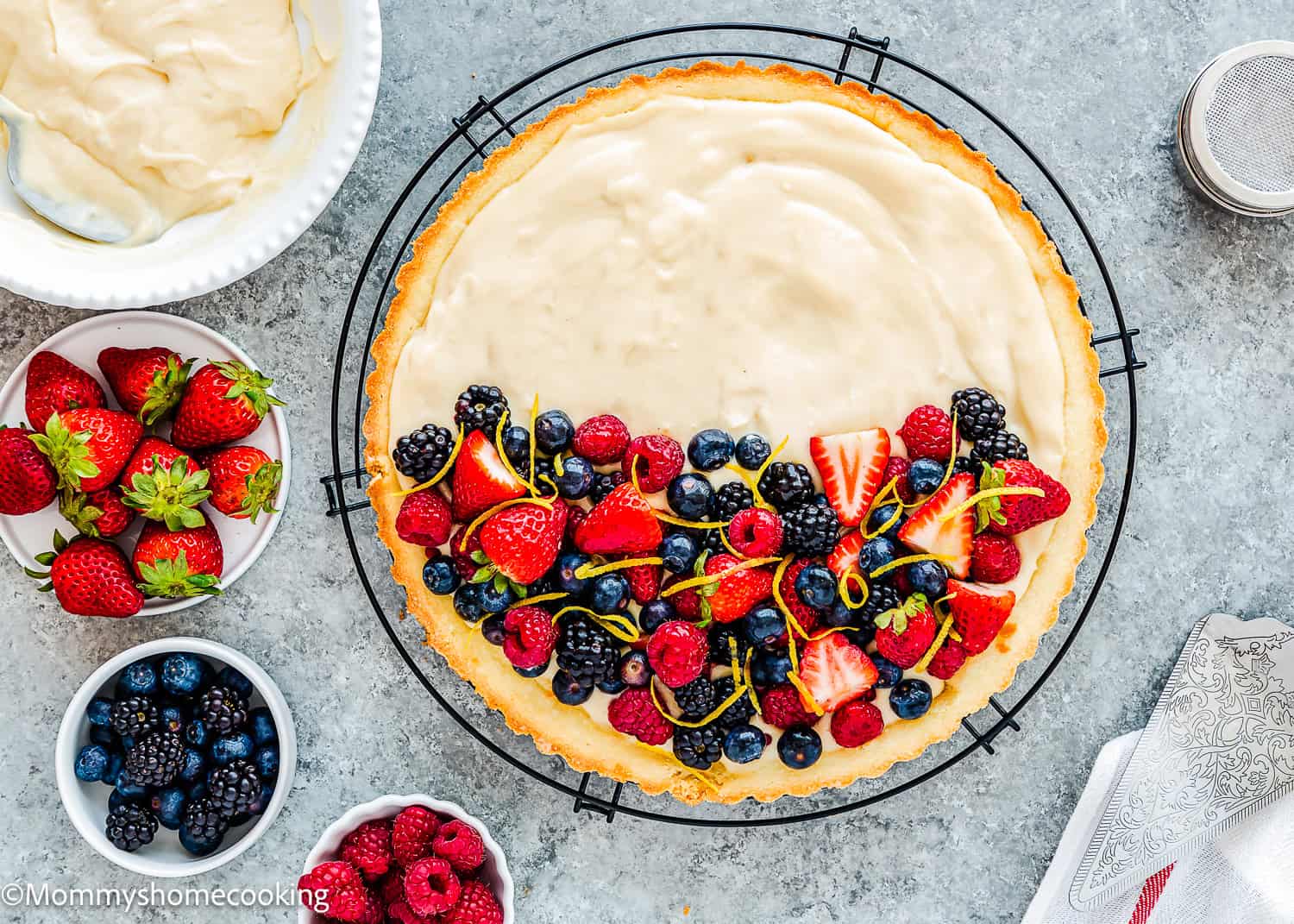 A partially decorated Easy Eggless Fruit Tart with Vanilla Pastry Cream, topped with assorted fresh berries and citrus zest, sits on a cooling rack. Bowls of strawberries, raspberries, and blackberries are nearby.