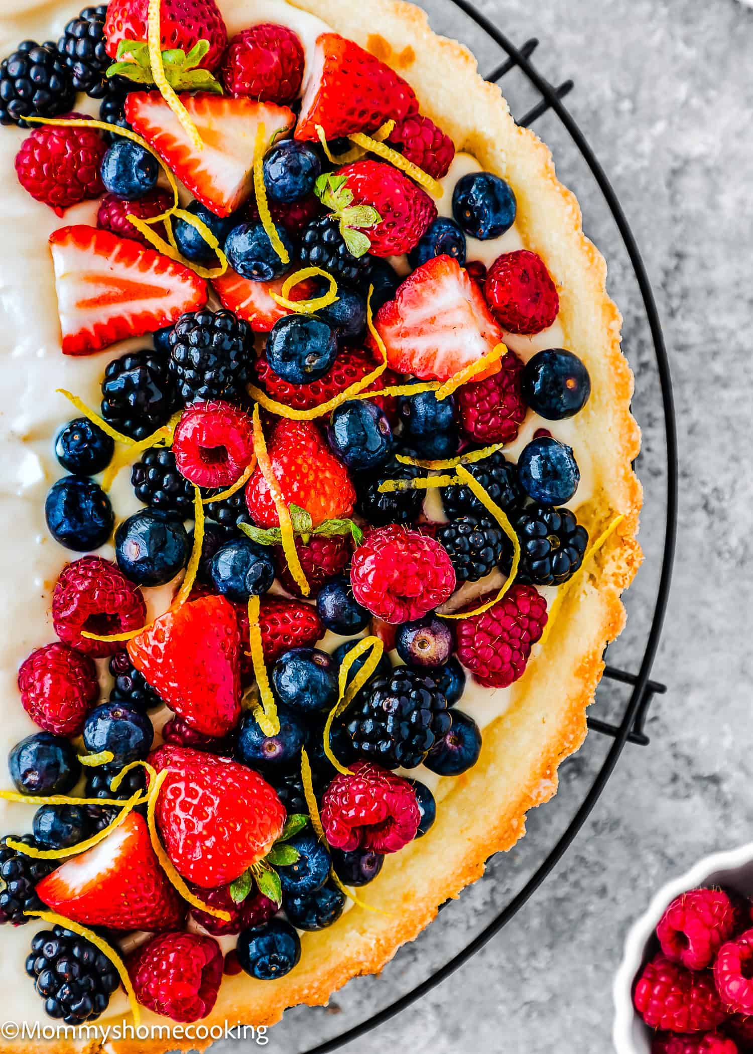 An Easy Eggless Fruit Tart with Vanilla Pastry Cream sits on a cooling rack, its creamy filling topped with strawberries, raspberries, blueberries, blackberries, and a sprinkle of lemon zest. Beside it rests a small bowl of fresh raspberries.