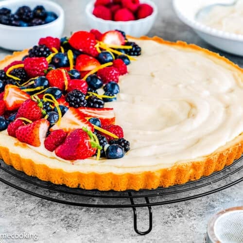 An Easy Eggless Fruit Tart with Vanilla Pastry Cream sits on a cooling rack, its creamy filling half topped with fresh strawberries, raspberries, blueberries, and lemon zest. In the background, bowls of fruit and cream add to the inviting scene.