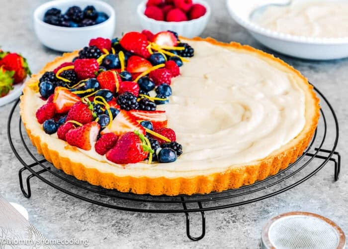 An Easy Eggless Fruit Tart with Vanilla Pastry Cream sits on a cooling rack, its creamy filling half topped with fresh strawberries, raspberries, blueberries, and lemon zest. In the background, bowls of fruit and cream add to the inviting scene.