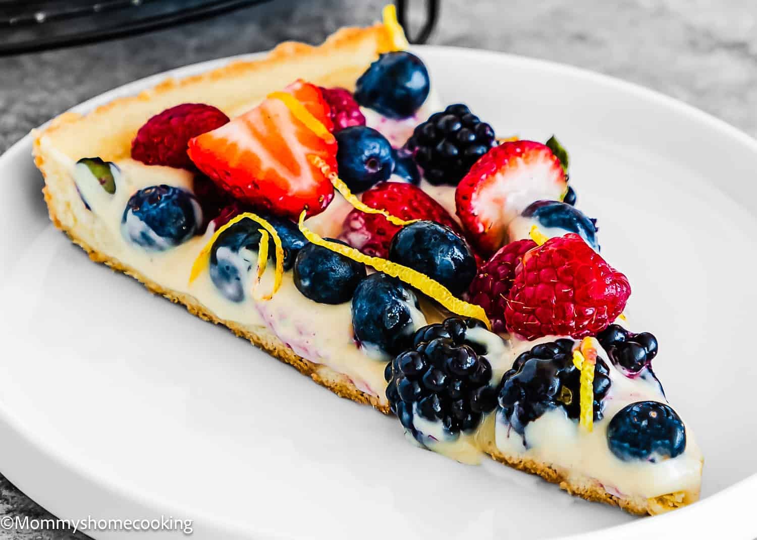 A slice of Easy Eggless Fruit Tart with Vanilla Pastry Cream is topped with strawberries, blueberries, blackberries, raspberries, lemon zest, and cream on a white plate.