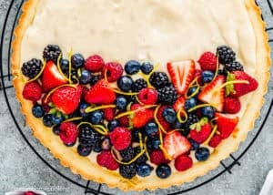 Half of an Easy Eggless Fruit Tart topped with strawberries, blueberries, raspberries, blackberries, and lemon zest sits on a cooling rack, with a creamy filling and a golden crust visible.