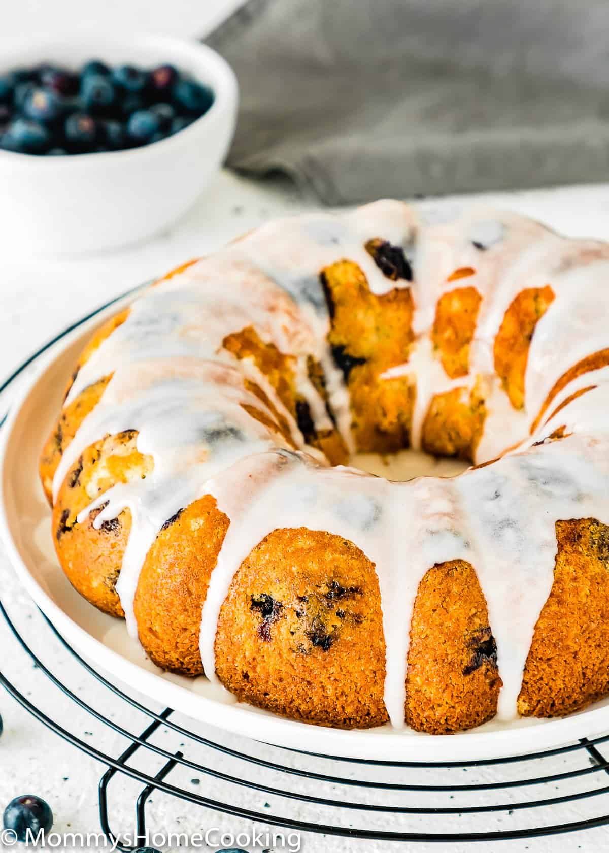 Eggless Lemon Blueberry bundt Cake on a plate with a bowl of blueberries in the backgraound.