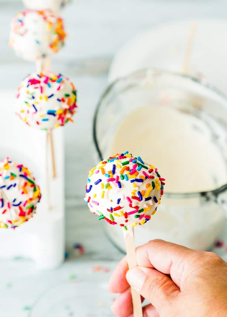 a hand holding an eggless cake pop being covered with melted candy melts and sprinkles.