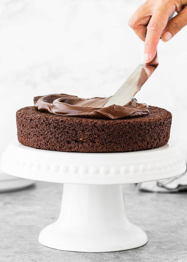 a spatula spreading chocolate ganache an egg-free chocolate cake layer over a cake stand. 