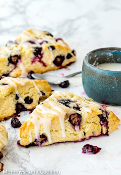 Eggless Blueberry Scones drizzled with lemon glaze over a marble surface with a small bowl on the background.