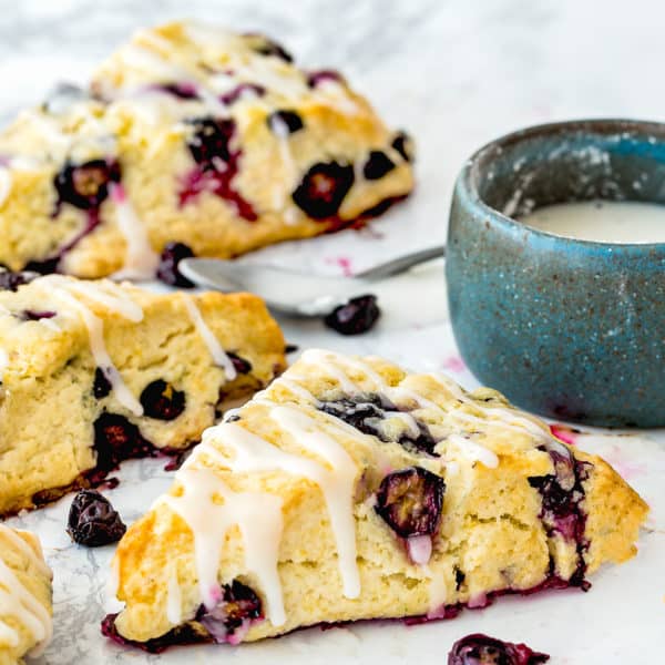 Eggless Blueberry Scones drizzled with lemon glaze over a marble surface with a small bowl on the background.